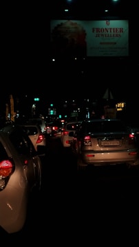 A nighttime traffic scene filled with closely packed cars, their red taillights illuminated. A large billboard for a jeweler is visible above the traffic, dimly lit by surrounding streetlights.