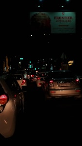 A nighttime traffic scene filled with closely packed cars, their red taillights illuminated. A large billboard for a jeweler is visible above the traffic, dimly lit by surrounding streetlights.
