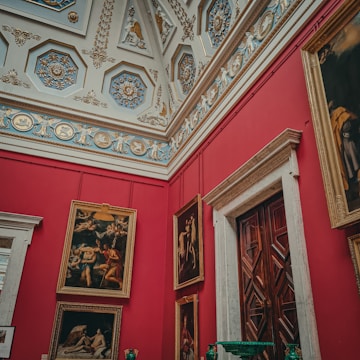 An ornate room with a richly decorated ceiling features intricate patterns and relief sculptures. The walls are vivid red, displaying several large framed paintings in classic gold frames. The door has a dark wood finish with geometric patterns and is framed by detailed molding.