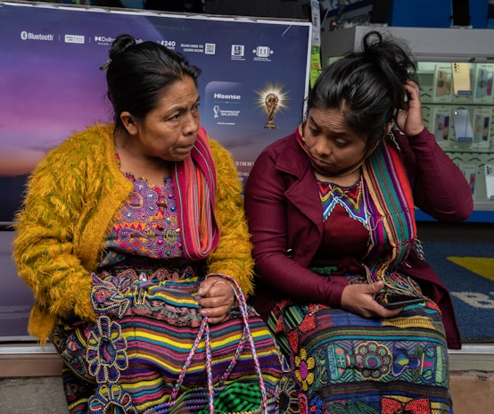 Two women sit closely together, dressed in vibrant traditional clothing featuring intricate patterns. One woman wears a mustard yellow sweater, while the other sports a burgundy jacket. Both have colorful woven textiles and appear engaged in conversation. Behind them, a display with electronic devices is partially visible.