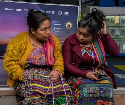 Two women sit closely together, dressed in vibrant traditional clothing featuring intricate patterns. One woman wears a mustard yellow sweater, while the other sports a burgundy jacket. Both have colorful woven textiles and appear engaged in conversation. Behind them, a display with electronic devices is partially visible.