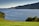 A serene coastal scene featuring multiple circular fish farming enclosures in a calm blue sea. The background consists of lush green hills and a distant island. The foreground includes a vibrant green grassy area with a few trees.