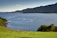 A serene coastal scene featuring multiple circular fish farming enclosures in a calm blue sea. The background consists of lush green hills and a distant island. The foreground includes a vibrant green grassy area with a few trees.