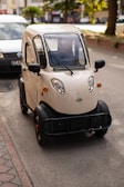 A compact, beige three-wheeled vehicle is parked on a city street. The design features rounded edges and a small windshield equipped with wipers. It has black trim on the wheel arches and bumper, and there are visible orange accents on the wheels. A white van is parked nearby, and trees can be seen in the background.