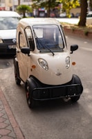 A compact, beige three-wheeled vehicle is parked on a city street. The design features rounded edges and a small windshield equipped with wipers. It has black trim on the wheel arches and bumper, and there are visible orange accents on the wheels. A white van is parked nearby, and trees can be seen in the background.