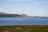 A serene coastal landscape with a vast blue sea stretching out towards distant hills. In the water, a series of rectangular fish farms or similar structures are present, with a small boat nearby. The foreground features a rocky, grassy area with sparse vegetation.