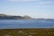 A serene coastal landscape with a vast blue sea stretching out towards distant hills. In the water, a series of rectangular fish farms or similar structures are present, with a small boat nearby. The foreground features a rocky, grassy area with sparse vegetation.