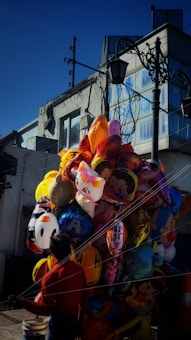 A street vendor is handling a large bunch of colorful helium balloons in various shapes and designs, including cartoon characters and emojis. The balloons are tightly grouped and create a vibrant contrast against the urban backdrop of a modern building with reflective glass windows. The vendor is wearing a red shirt and is mostly in shadow, adding a sense of mystery to the scene.