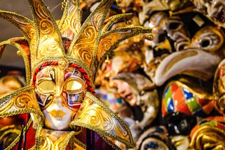 A collection of ornate and colorful Venetian masks, with one prominent gold and red jester mask in the foreground. The masks feature intricate details and a variety of designs, showcasing craftsmanship typical of carnival celebrations.
