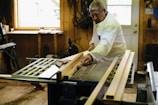 An older man wearing protective glasses and a white coat is working on a piece of wood using a table saw. The workshop is well-lit by a window nearby, and there are several tools and pieces of wood scattered around. The walls are wooden, contributing to the rustic feel of the workshop.