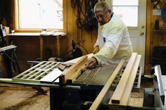 An older man wearing protective glasses and a white coat is working on a piece of wood using a table saw. The workshop is well-lit by a window nearby, and there are several tools and pieces of wood scattered around. The walls are wooden, contributing to the rustic feel of the workshop.