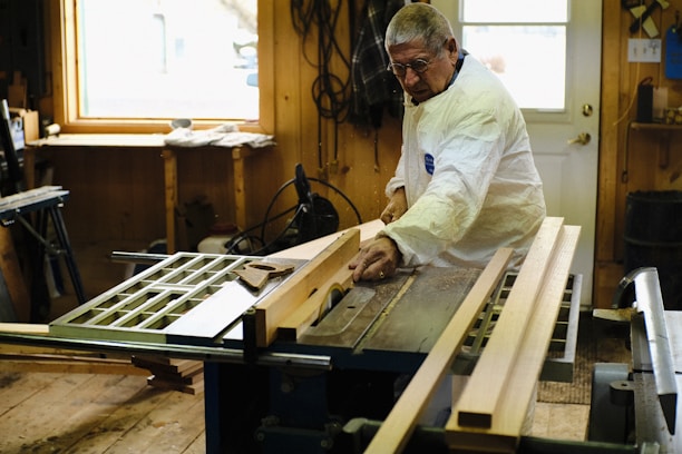 An older man wearing protective glasses and a white coat is working on a piece of wood using a table saw. The workshop is well-lit by a window nearby, and there are several tools and pieces of wood scattered around. The walls are wooden, contributing to the rustic feel of the workshop.