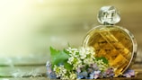 A round, diamond-patterned perfume bottle sits beside a cluster of delicate white and purple flowers on a wooden surface. The background is softly blurred, creating a serene and natural ambiance.