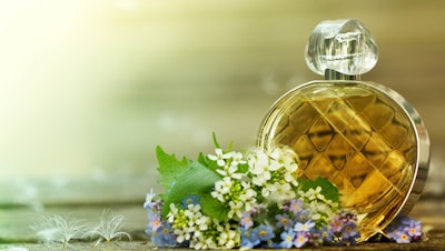 A round, diamond-patterned perfume bottle sits beside a cluster of delicate white and purple flowers on a wooden surface. The background is softly blurred, creating a serene and natural ambiance.