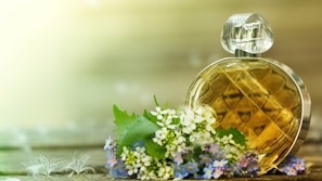 A round, diamond-patterned perfume bottle sits beside a cluster of delicate white and purple flowers on a wooden surface. The background is softly blurred, creating a serene and natural ambiance.