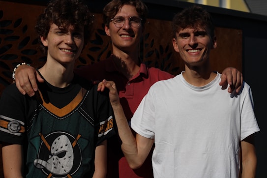 Three young males are standing closely together, smiling at the camera. One is wearing a t-shirt with a sports logo, another is in a red polo shirt, and the third is in a white t-shirt. Their arms are around each other's shoulders in a gesture of camaraderie.