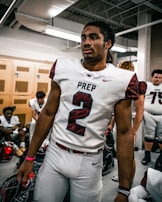 A football player wearing the number 2 jersey stands in a locker room, holding a helmet. He appears focused and ready, surrounded by teammates who are sitting on benches, also wearing football gear. The room has lockers and a ceiling with exposed pipes.