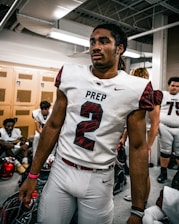 A football player wearing the number 2 jersey stands in a locker room, holding a helmet. He appears focused and ready, surrounded by teammates who are sitting on benches, also wearing football gear. The room has lockers and a ceiling with exposed pipes.