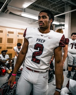 A football player wearing the number 2 jersey stands in a locker room, holding a helmet. He appears focused and ready, surrounded by teammates who are sitting on benches, also wearing football gear. The room has lockers and a ceiling with exposed pipes.