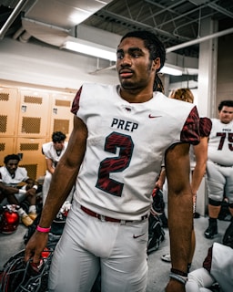 A football player wearing the number 2 jersey stands in a locker room, holding a helmet. He appears focused and ready, surrounded by teammates who are sitting on benches, also wearing football gear. The room has lockers and a ceiling with exposed pipes.