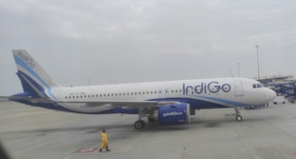 An airplane with the IndiGo logo is stationed on the tarmac. The aircraft is painted primarily in white and blue colors. A person wearing a yellow outfit walks nearby, and the sky is overcast with clouds.