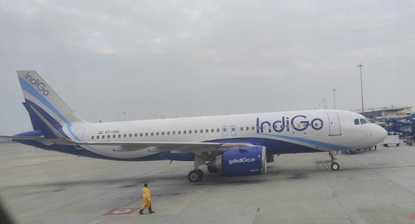 An airplane with the IndiGo logo is stationed on the tarmac. The aircraft is painted primarily in white and blue colors. A person wearing a yellow outfit walks nearby, and the sky is overcast with clouds.