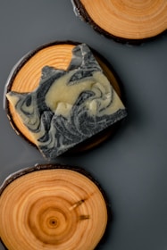 A piece of intricately swirled soap rests on a circular wooden coaster, which displays natural wood grain patterns. Another wooden coaster is partially visible, providing a rustic and aesthetically pleasing composition.