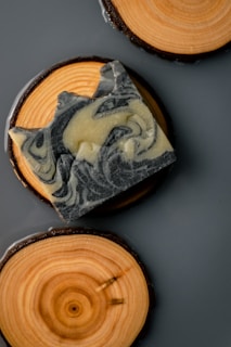 A piece of intricately swirled soap rests on a circular wooden coaster, which displays natural wood grain patterns. Another wooden coaster is partially visible, providing a rustic and aesthetically pleasing composition.