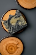 A piece of intricately swirled soap rests on a circular wooden coaster, which displays natural wood grain patterns. Another wooden coaster is partially visible, providing a rustic and aesthetically pleasing composition.