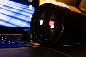 A close-up of a camera lens resting on a laptop keyboard. The lens is prominently featured in the foreground, showing its brand and specifications. The laptop screen displays a blue and white abstract pattern, with some bokeh lights reflecting off the lens. The scene is dimly lit, creating a focused and professional atmosphere.