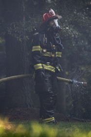 A firefighter in full protective gear, including a helmet and mask, stands holding a hose spraying water. They are surrounded by a misty environment with trees in the background.