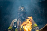 A firefighter wearing protective gear and a helmet is surrounded by smoke and flames, with sparks floating in the air.