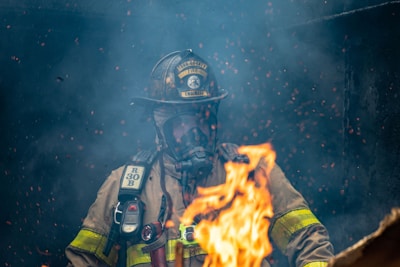 A firefighter wearing protective gear and a helmet is surrounded by smoke and flames, with sparks floating in the air.