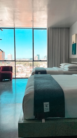 A modern hotel room with two neatly made beds, each with a dark blanket at the foot. The room features a large floor-to-ceiling window offering a view of a cityscape with various buildings under a clear blue sky. A red chair is positioned near the window, and the space is illuminated with natural light.