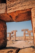 Ancient stone structures arranged in a doorway-like formation stand amidst a barren landscape, with a clear blue sky overhead. The formations are made of large, weathered blocks, hinting at a historical or archaeological site.