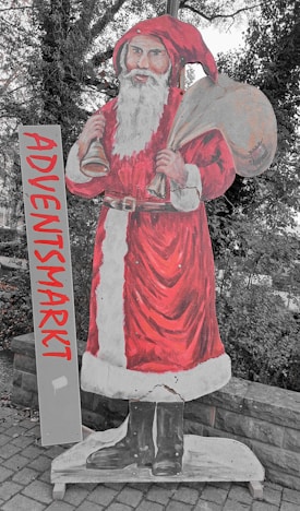 A painted cutout of Santa Claus stands outdoors, holding a sack over his shoulder and a bell in one hand. He is dressed in a traditional red and white outfit, complete with a fluffy white beard and black boots. Beside him is a vertical sign with the word 'ADVENTSMARKT' written in bold red letters. The scene is set against a backdrop of trees and a cobblestone path.