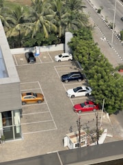 An aerial view of a parking lot with multiple cars parked in designated spaces. The lot is surrounded by palm trees and greenery, with a paved road running alongside. Cars of different colors, including white, red, blue, and orange, are visible. A building with glass windows is adjacent to the parking area.