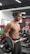 A muscular man with long hair tied in a bun is lifting weights in a gym. He is shirtless and wearing black athletic pants. The gym background includes equipment like weights and benches, with a vivid mural on the wall depicting a superhero.