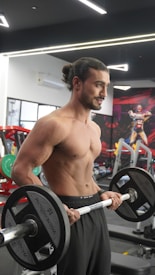 A muscular man with long hair tied in a bun is lifting weights in a gym. He is shirtless and wearing black athletic pants. The gym background includes equipment like weights and benches, with a vivid mural on the wall depicting a superhero.