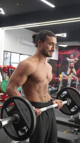 A muscular man with long hair tied in a bun is lifting weights in a gym. He is shirtless and wearing black athletic pants. The gym background includes equipment like weights and benches, with a vivid mural on the wall depicting a superhero.