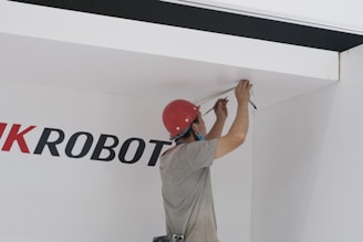 A person wearing a red hard hat and a face mask is working on the ceiling, possibly installing or repairing electrical wiring. The individual is holding tools in their hands and is focused on the task. The wall behind them has partially visible text in large black and red letters.