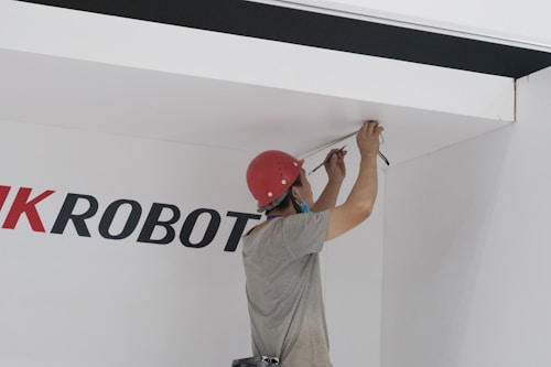 A person wearing a red hard hat and a face mask is working on the ceiling, possibly installing or repairing electrical wiring. The individual is holding tools in their hands and is focused on the task. The wall behind them has partially visible text in large black and red letters.