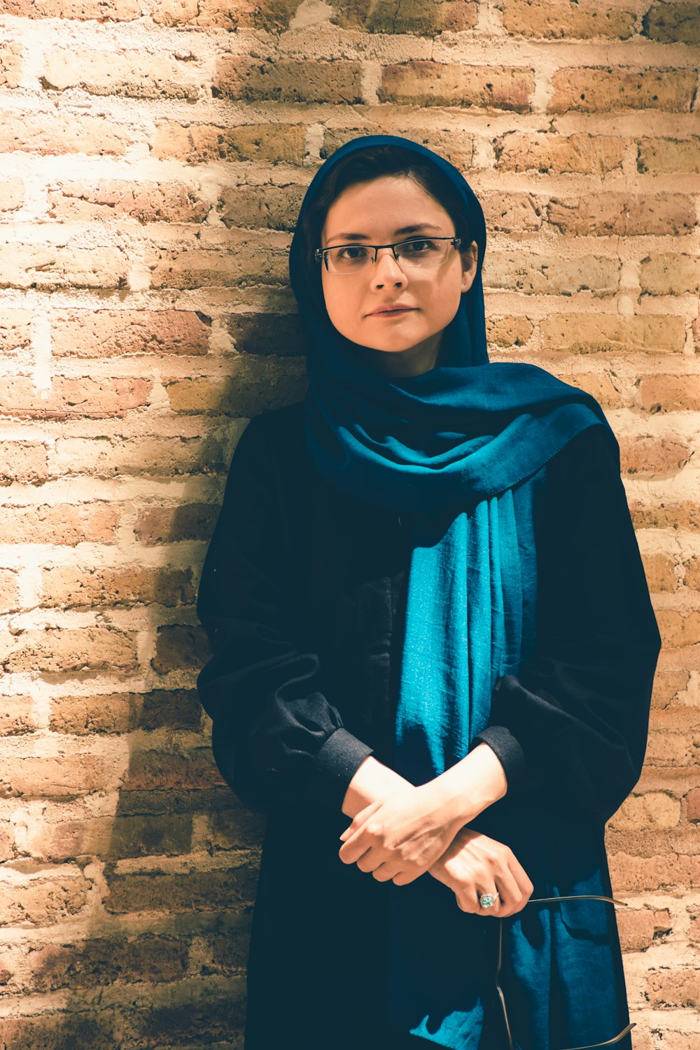 a woman in a black scarf and glasses standing in front of a brick wall