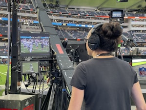 A person with headphones is operating a large camera setup at a sports stadium. The camera equipment is focused on a screen showing a live sports event taking place on a football field. The stadium is filled with spectators, and the atmosphere is busy with technical setup and monitors.