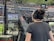 A person with headphones is operating a large camera setup at a sports stadium. The camera equipment is focused on a screen showing a live sports event taking place on a football field. The stadium is filled with spectators, and the atmosphere is busy with technical setup and monitors.