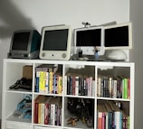 A collection of vintage computers and monitors displayed on a white shelving unit, accompanied by a variety of books neatly organized on the shelves. The top shelf features retro Apple computers and gadgets, including a webcam and a small figure. Below, books of different genres and topics line the shelves, with a few gaming controllers and a model motorcycle in the lower compartments.