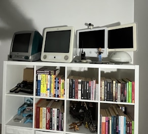 A collection of vintage computers and monitors displayed on a white shelving unit, accompanied by a variety of books neatly organized on the shelves. The top shelf features retro Apple computers and gadgets, including a webcam and a small figure. Below, books of different genres and topics line the shelves, with a few gaming controllers and a model motorcycle in the lower compartments.