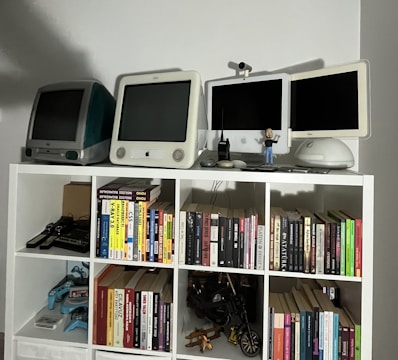 A collection of vintage computers and monitors displayed on a white shelving unit, accompanied by a variety of books neatly organized on the shelves. The top shelf features retro Apple computers and gadgets, including a webcam and a small figure. Below, books of different genres and topics line the shelves, with a few gaming controllers and a model motorcycle in the lower compartments.