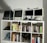 A collection of vintage computers and monitors displayed on a white shelving unit, accompanied by a variety of books neatly organized on the shelves. The top shelf features retro Apple computers and gadgets, including a webcam and a small figure. Below, books of different genres and topics line the shelves, with a few gaming controllers and a model motorcycle in the lower compartments.