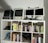 A collection of vintage computers and monitors displayed on a white shelving unit, accompanied by a variety of books neatly organized on the shelves. The top shelf features retro Apple computers and gadgets, including a webcam and a small figure. Below, books of different genres and topics line the shelves, with a few gaming controllers and a model motorcycle in the lower compartments.